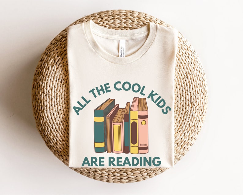 Puede incluir: Una camiseta beige con el texto "All the cool kids are reading" en verde y marr&oacute;n. Una pila de libros coloridos est&aacute; ilustrada en el centro de la camiseta.