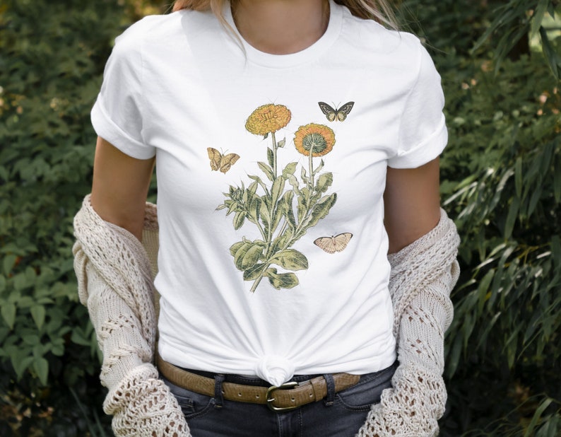 May include: White t-shirt with a botanical illustration of yellow flowers and butterflies. The flowers have a vintage style and are surrounded by green leaves.