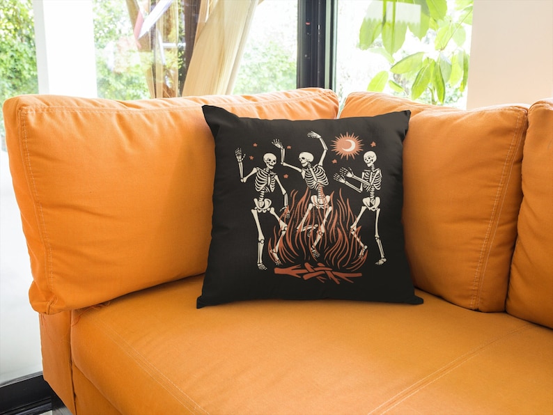 May include: A black throw pillow with a design of three skeletons dancing around a fire. The skeletons are white and the fire is orange. There are stars and a crescent moon in the background.