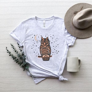 May include: A gray t-shirt with a black and orange owl graphic. The owl is perched on a branch with a crescent moon and stars in the background.