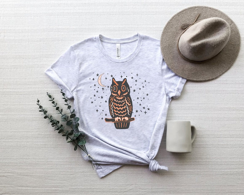May include: A gray t-shirt with a black and orange owl graphic. The owl is perched on a branch with a crescent moon and stars in the background.