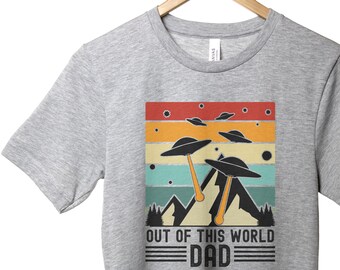 Puede incluir: Camiseta gris con un gráfico retro de platillos voladores y montañas. El texto "Out of this world Dad" está impreso debajo del gráfico.