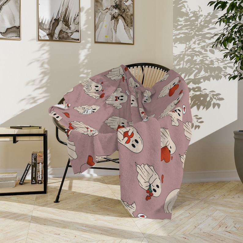 May include: A pink throw blanket with a repeating pattern of white ghosts holding red hearts. The blanket is draped over a wicker chair.
