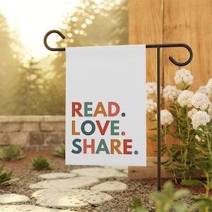 May include: A white garden flag with the text "READ. LOVE. SHARE." in colorful, stylized letters. The flag is attached to a black metal garden flag stand.