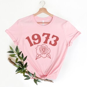 May include: A pink t-shirt with a red 1973 and a rose graphic.