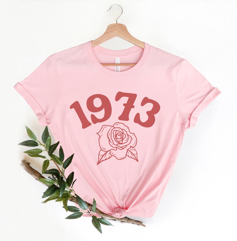 May include: A pink t-shirt with a red 1973 and a rose graphic.