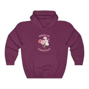 Peut inclure: Un sweat &agrave; capuche bordeaux avec une poche blanche et le texte "WHAT IN CARNATION?" en lettres roses. Un bouquet de &oelig;illets roses et blancs est imprim&eacute; sous le texte.
