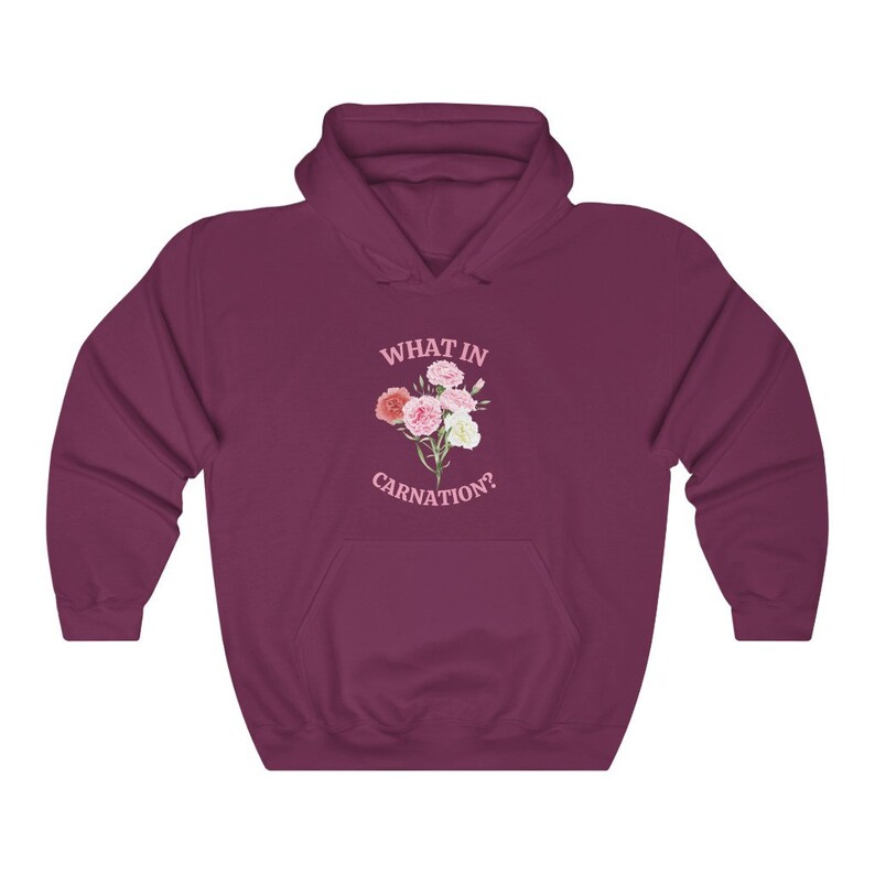 Peut inclure: Un sweat &agrave; capuche bordeaux avec une poche blanche et le texte "WHAT IN CARNATION?" en lettres roses. Un bouquet de &oelig;illets roses et blancs est imprim&eacute; sous le texte.