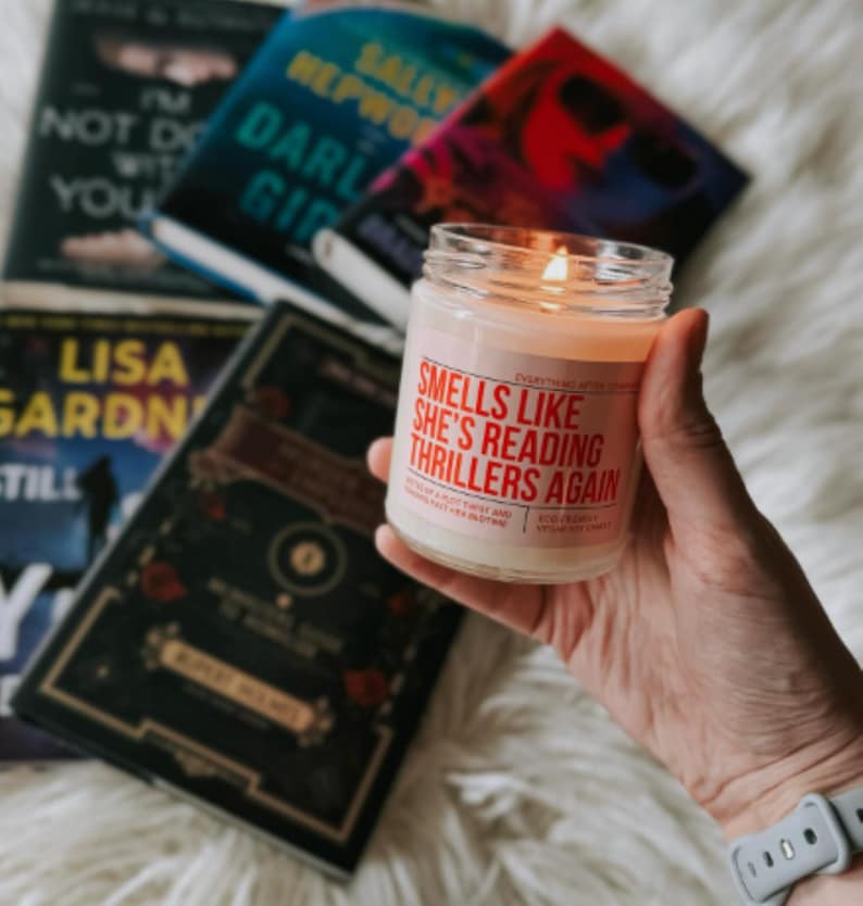 May include: A white candle with a red label that reads "Smells Like She's Reading Thrillers Again". The candle is sitting on a table with several books.