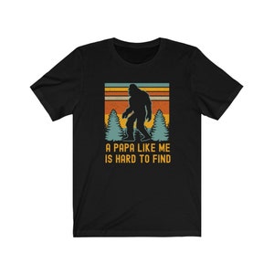 May include: Black t-shirt with a retro-style graphic of a Bigfoot walking through a forest. The text "A Papa Like Me Is Hard To Find" is printed below the graphic.