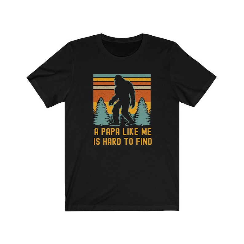 May include: Black t-shirt with a retro-style graphic of a Bigfoot walking through a forest. The text "A Papa Like Me Is Hard To Find" is printed below the graphic.