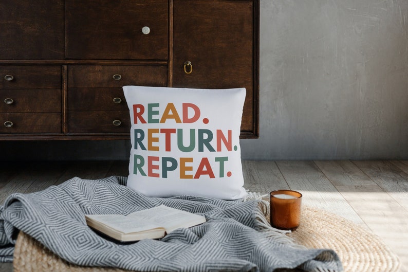 May include: White throw pillow with the text "READ. RETURN. REPEAT." in colorful letters. The pillow is on a gray and white blanket with a book and a candle.