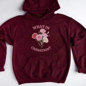 Peut inclure: Un sweat &agrave; capuche bordeaux avec une poche blanche et un motif rose qui dit "What in Carnation?" avec un bouquet de &oelig;illets roses.