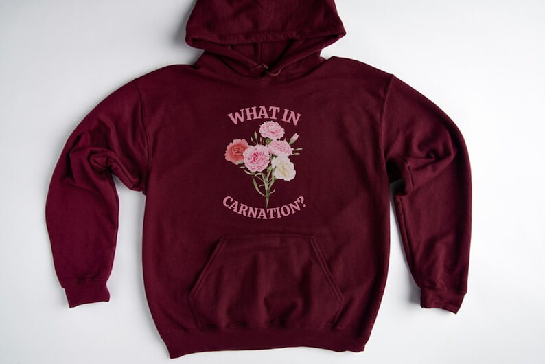 Peut inclure: Un sweat &agrave; capuche bordeaux avec une poche blanche et un motif rose qui dit "What in Carnation?" avec un bouquet de &oelig;illets roses.