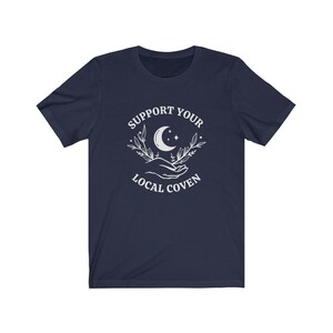 May include: Navy blue t-shirt with a white graphic design featuring a crescent moon, stars, and two hands holding a sprig of leaves. The text "Support Your Local Coven" is printed below the graphic.