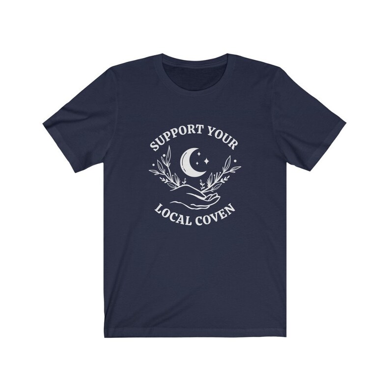 May include: Navy blue t-shirt with a white graphic design featuring a crescent moon, stars, and two hands holding a sprig of leaves. The text "Support Your Local Coven" is printed below the graphic.