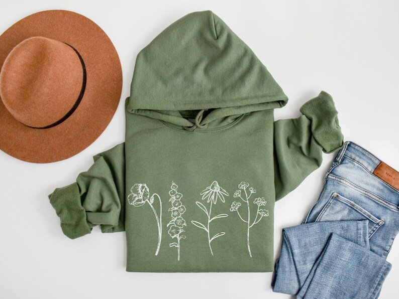 May include: A green hooded sweatshirt with a white outline of various flowers on the front. The sweatshirt is laid flat on a white surface with a brown hat and blue jeans beside it.