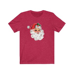 May include: A red t-shirt with a vintage illustration of Santa Claus's face. The illustration is in a cartoon style with a red hat, white beard, and a jolly expression.