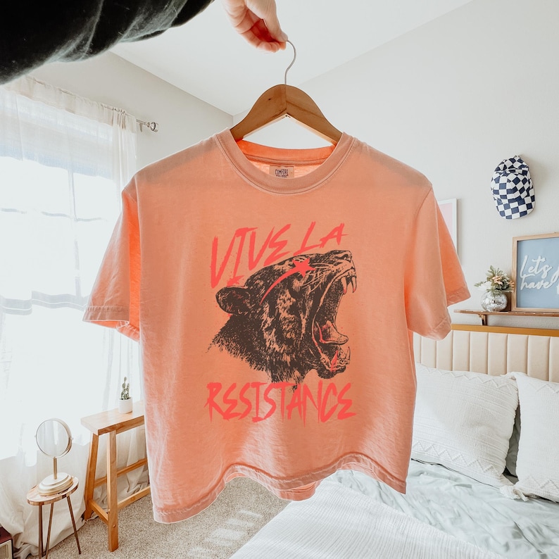 May include: A peach-colored cropped t-shirt with a graphic of a roaring tiger and the text "Vive La Resistance" in red.