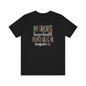 Op de afbeelding: Zwart T-shirt met de tekst "MY FAVORITE baseball PLAYER CALLS ME mom" in kleurrijke, speelse letters. Een honkbalafbeelding is inbegrepen. Het shirt is gemaakt van een zacht materiaal.