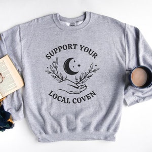 May include: Gray sweatshirt with the text "Support Your Local Coven" in black lettering. The design includes a crescent moon, stars, and two hands holding the moon.