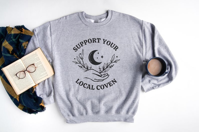 May include: Gray sweatshirt with the text "Support Your Local Coven" in black lettering. The design includes a crescent moon, stars, and two hands holding the moon.