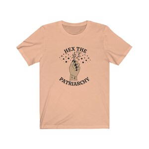 May include: Peach-colored t-shirt with the text "HEX THE PATRIARCHY" above an illustration of a hand holding a sprig of leaves. The hand has black nail polish and a ring. Stars and a crescent moon are above the hand.