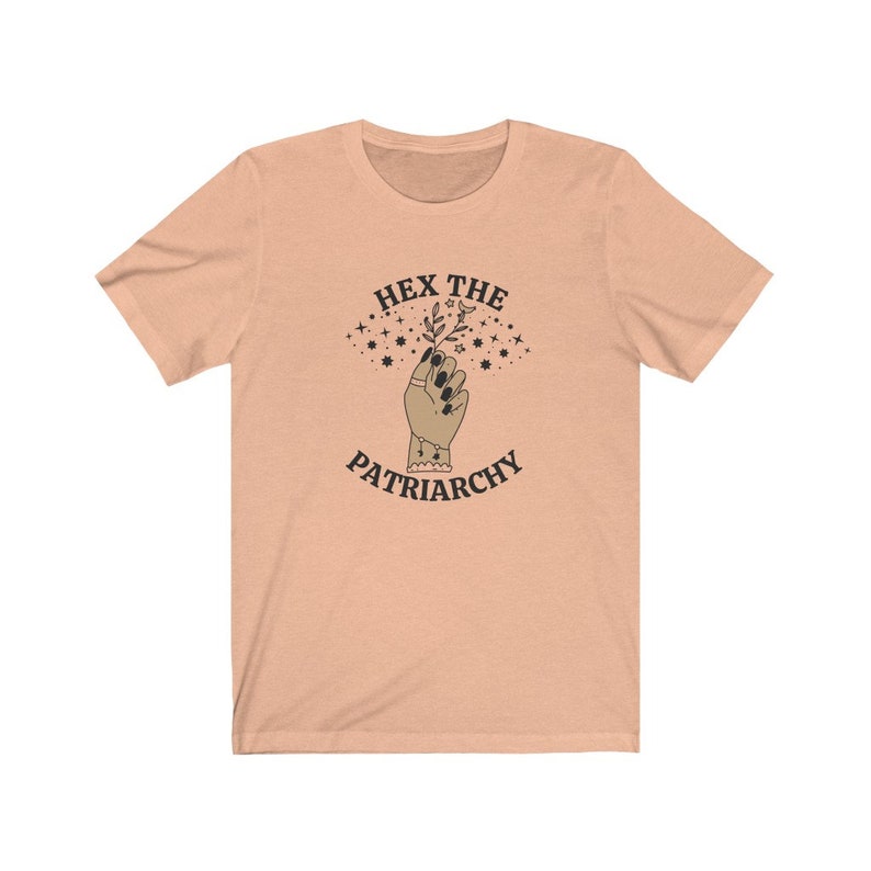 May include: Peach-colored t-shirt with the text "HEX THE PATRIARCHY" above an illustration of a hand holding a sprig of leaves. The hand has black nail polish and a ring. Stars and a crescent moon are above the hand.