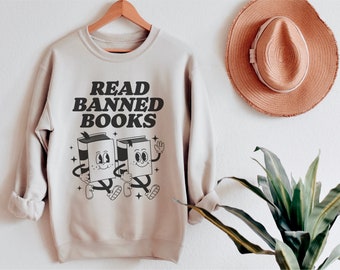 May include: A beige sweatshirt with the text "READ BANNED BOOKS" in black lettering. Two cartoon book characters are walking side-by-side, holding hands.