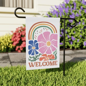 May include: A white garden flag with a floral design and the word "WELCOME" in orange. The design includes a rainbow, pink, blue, and orange flowers. The flag is displayed on a black metal stand in a grassy yard.