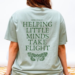 May include: A light green t-shirt with the text "INSPIRE, GROW, FLY!" and "HELPING LITTLE MINDS TAKE FLIGHT" in dark green. A detailed illustration of a moth is below the text. The shirt is a casual style.
