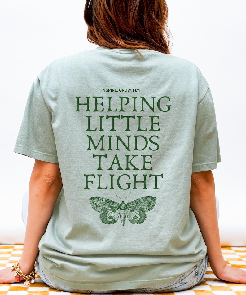 May include: A light green t-shirt with the text "INSPIRE, GROW, FLY!" and "HELPING LITTLE MINDS TAKE FLIGHT" in dark green. A detailed illustration of a moth is below the text. The shirt is a casual style.