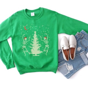 May include: Green Christmas sweater with two skeletons dancing around a Christmas tree. The sweater has a white crescent moon and snowflakes on it.