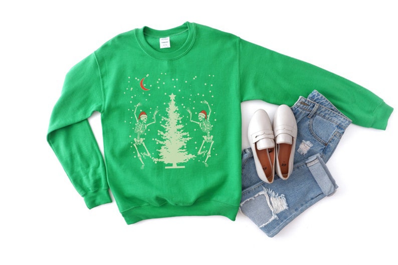 May include: Green Christmas sweater with two skeletons dancing around a Christmas tree. The sweater has a white crescent moon and snowflakes on it.