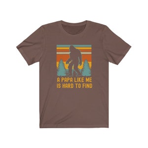May include: A brown t-shirt with a retro-style graphic of a Bigfoot walking through a forest. The text "A Papa Like Me Is Hard To Find" is printed below the graphic.