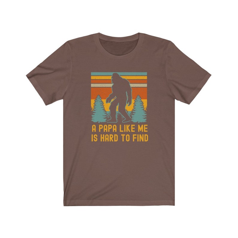 May include: A brown t-shirt with a retro-style graphic of a Bigfoot walking through a forest. The text "A Papa Like Me Is Hard To Find" is printed below the graphic.