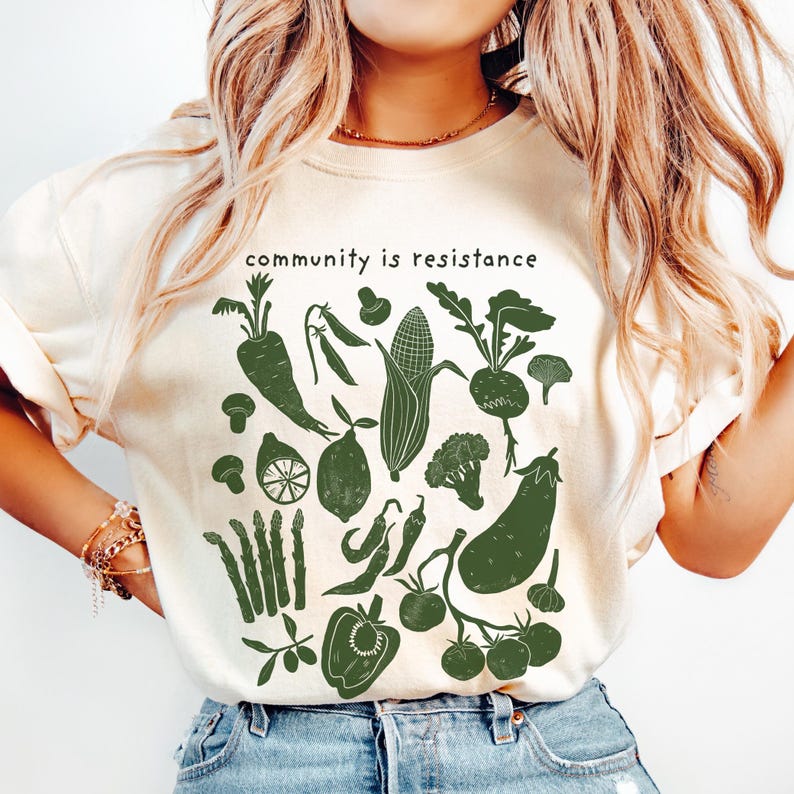 May include: Cream-colored t-shirt with a dark green illustration of various vegetables, including carrots, corn, and tomatoes. The text "community is resistance" is printed above the vegetables. The shirt is made of a soft material.