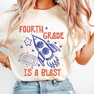 May include: A cream-colored short-sleeve t-shirt featuring a blue graphic of a rocket ship, a spiral galaxy, and a smiling star. The text "FOURTH GRADE IS A BLAST" is printed in red block lettering above and below the central space-themed illustration.