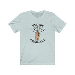 May include: Light blue t-shirt with the text "HEX THE PATRIARCHY" above an illustration of a hand holding a sprig of leaves and stars. The hand has black nail polish and a ring. The shirt is a short-sleeved crew neck.