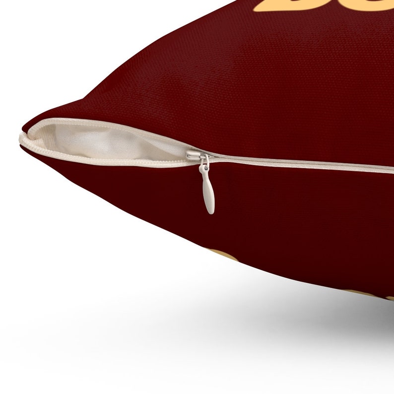 May include: A deep maroon throw pillow cover featuring a hidden white zip closure along the side seam. The fabric has a visible woven texture, and a portion of a light yellow graphic design is partially visible at the top edge of the pillow.