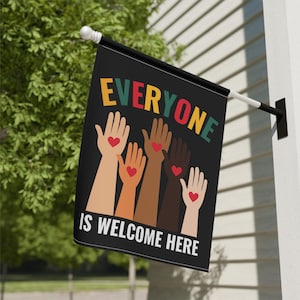 May include: A black flag with the words "Everyone is Welcome Here" in colorful letters. The flag features a design of five hands of different skin tones reaching up with red hearts in their palms.