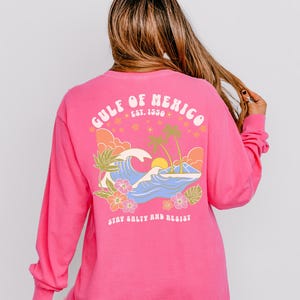 Puede incluir: Camiseta rosa de manga larga con un dise&ntilde;o gr&aacute;fico retro que presenta una ola, palmeras y el texto "Gulf of Mexico Est. 1550" y "Stay Salty and Resist".