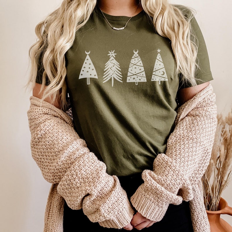 May include: A green t-shirt with a white graphic of five stylized Christmas trees. The trees have different designs, including stars, moons, and crescent moons. The person wearing the shirt is also wearing a beige cardigan.