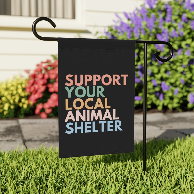 May include: A black garden flag with the text "SUPPORT YOUR LOCAL ANIMAL SHELTER" in colorful letters. The flag is displayed on a black metal garden flag stand.