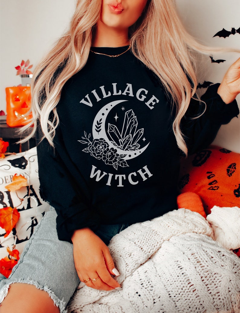 May include: A black sweatshirt with a white graphic design of a crescent moon with crystals and flowers. The text "Village Witch" is printed below the moon.
