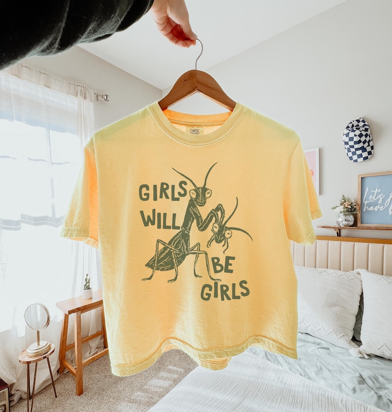 May include: Yellow t-shirt with the text "GIRLS WILL BE GIRLS" and a green illustration of two praying mantises. The shirt is hanging on a wooden hanger.