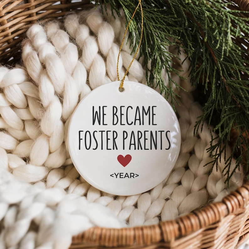 May include: A white ceramic ornament with the text "WE BECAME FOSTER PARENTS" in black, a red heart, and the word "<YEAR>" below. The ornament hangs from a gold string and is set against a white knitted blanket and greenery.