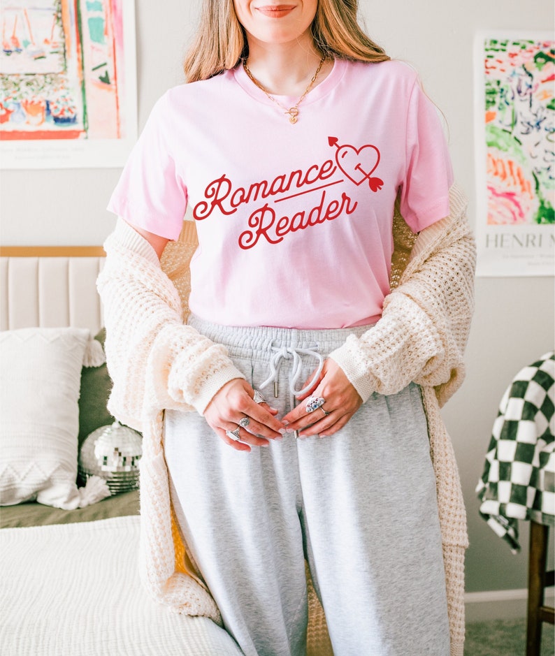 K&ouml;nnte beinhalten: Hellrosa T-Shirt mit rotem Schriftzug "Romance Reader" und einem Herz mit Pfeil. Dazu eine graue Jogginghose und eine cremefarbene Strickjacke.