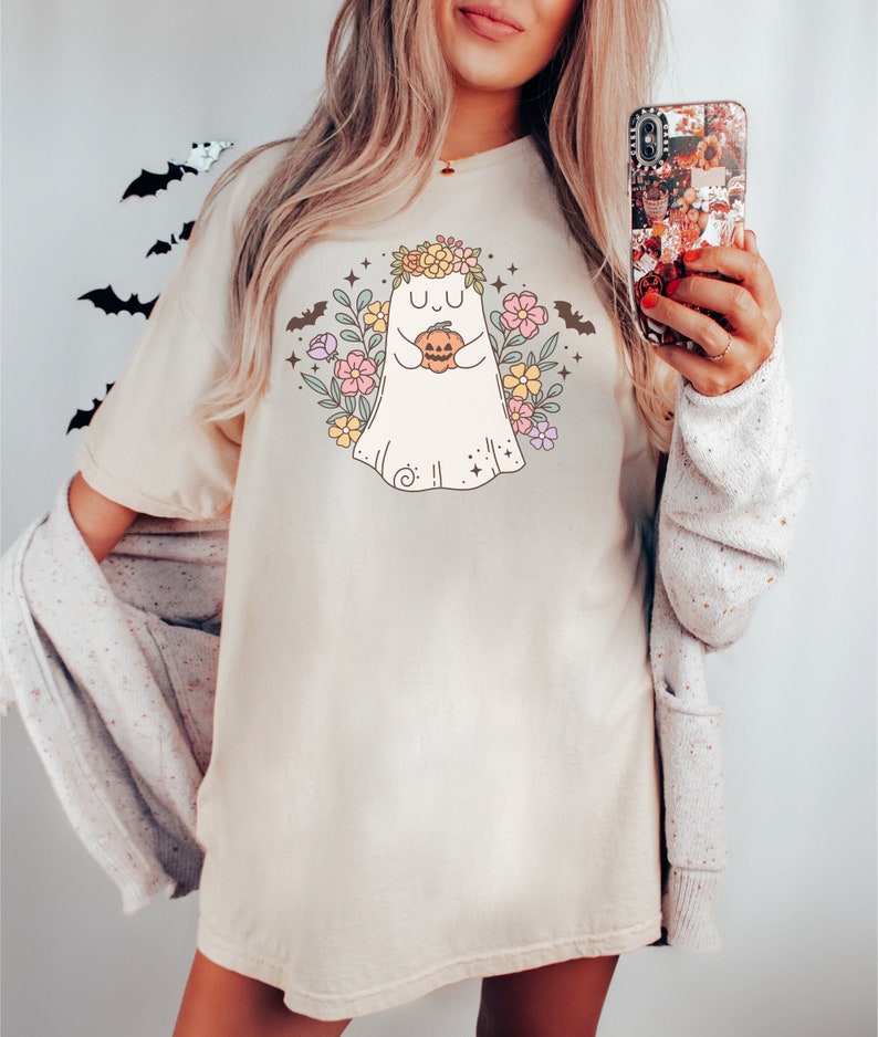 May include: A white t-shirt with a cute cartoon ghost wearing a flower crown and holding a pumpkin. The ghost is surrounded by flowers and bats.