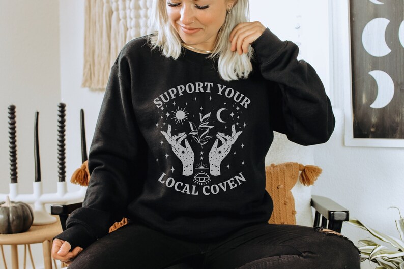 Puede incluir: Sudadera negra con un dise&ntilde;o gr&aacute;fico blanco que dice "Support Your Local Coven" con una luna creciente, un sol y dos manos sosteniendo una planta.